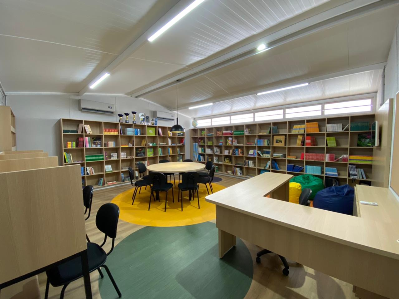 Biblioteca com estante de livros, mesa redonda com cadeiras, recepção e cabines individuais de estudo.