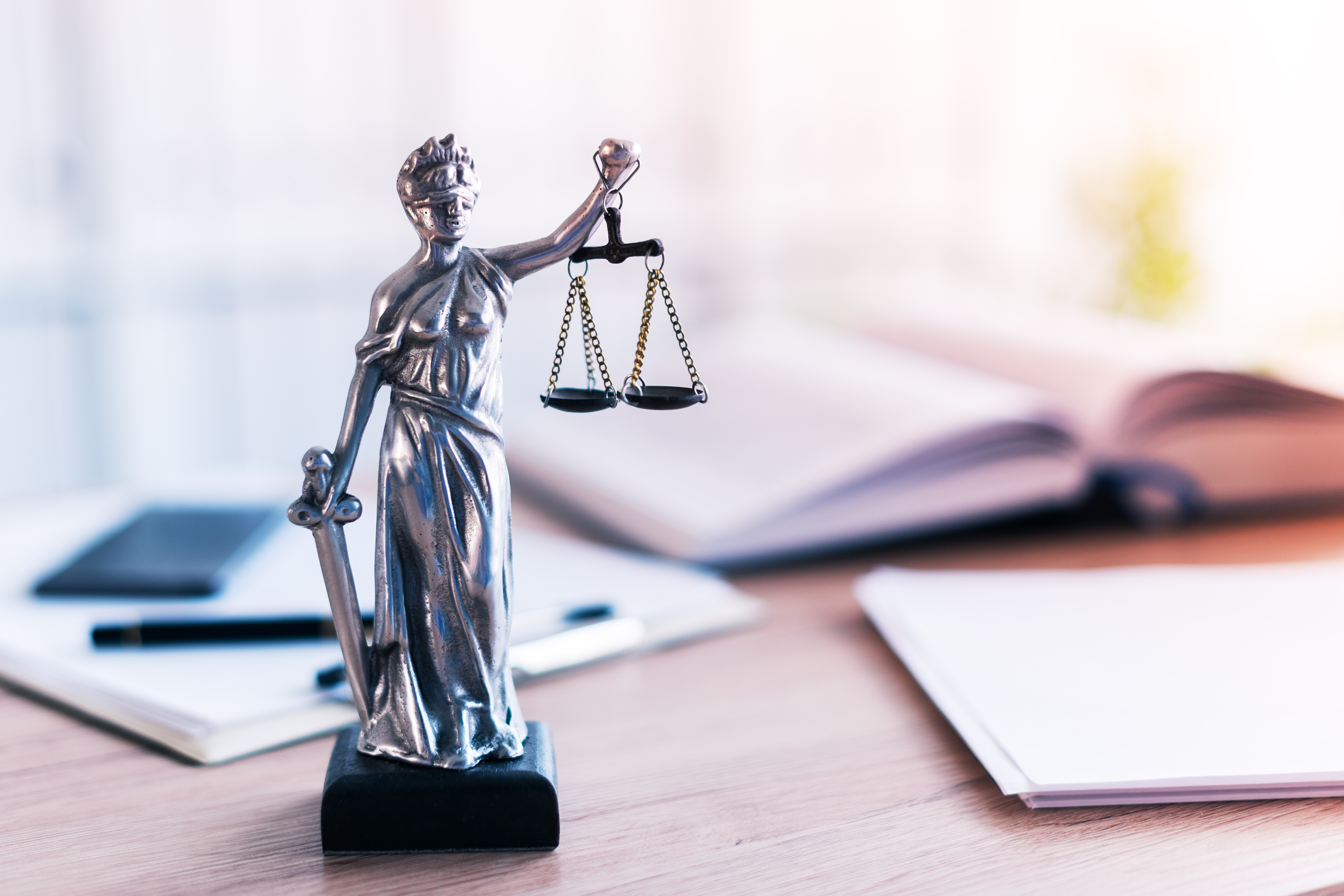 Descrição da Imagem: Estatua da Justiça sobre a mesa. Ao fundo, estão desfocados um livro aberto, papeis de anotação e uma caneta.