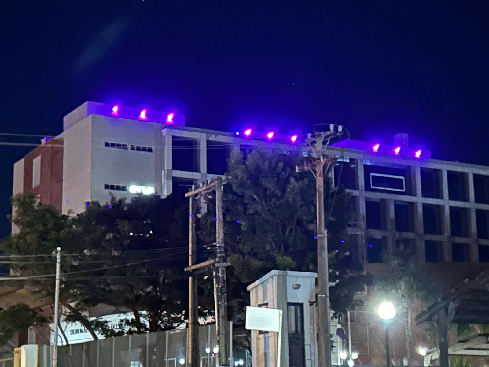 a Imagem mostra a fachada do prédio sede do TRT24 no período da noite. No terraço do prédio, lâmpadas iluminam a fachada com a cor roxa.