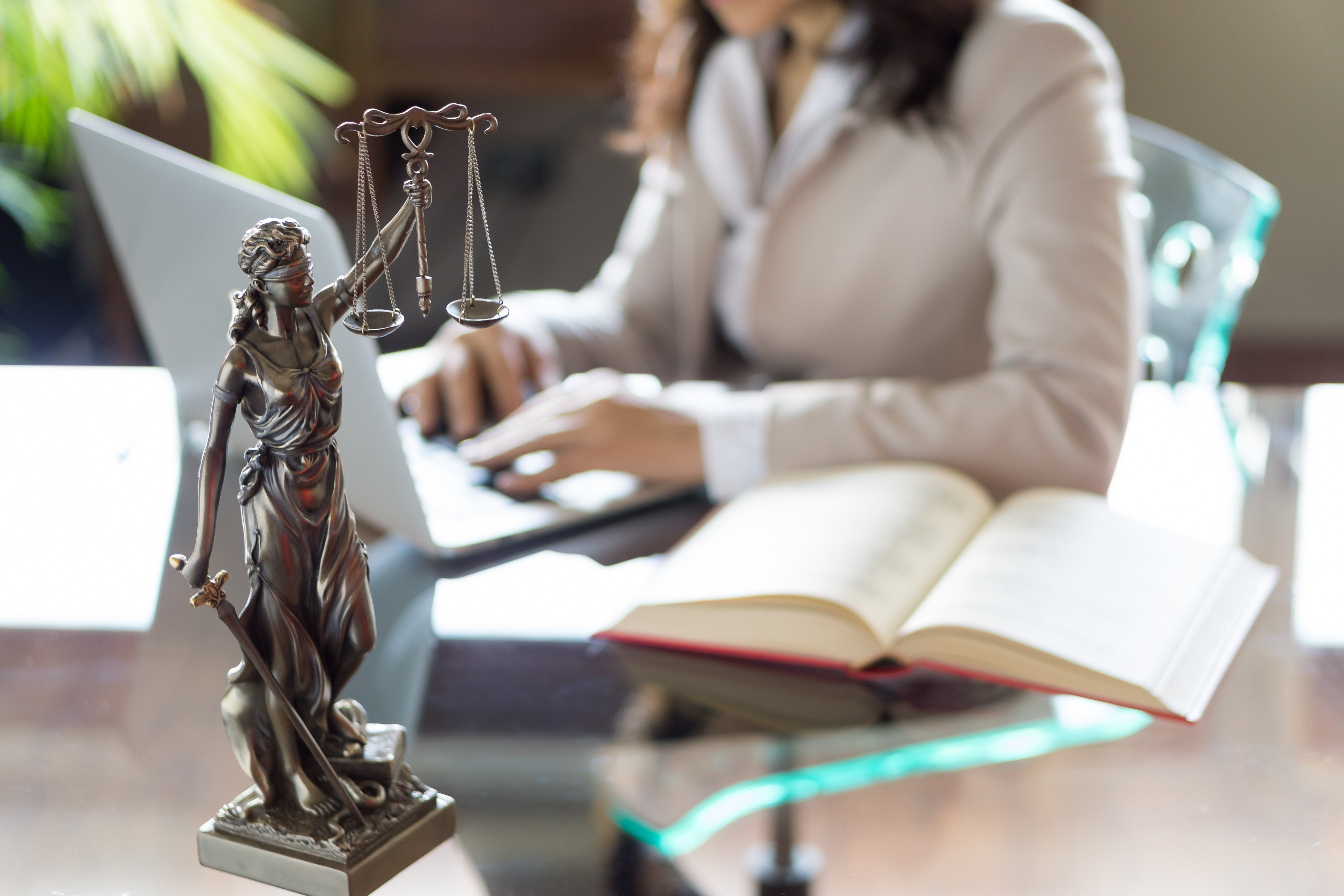 Imagem contendo estatueta da figura símbolo da justiça em cima de uma mesa com tampo de vidro. Ao fundo, à mesa, uma mulher utiliza um computador portátil.