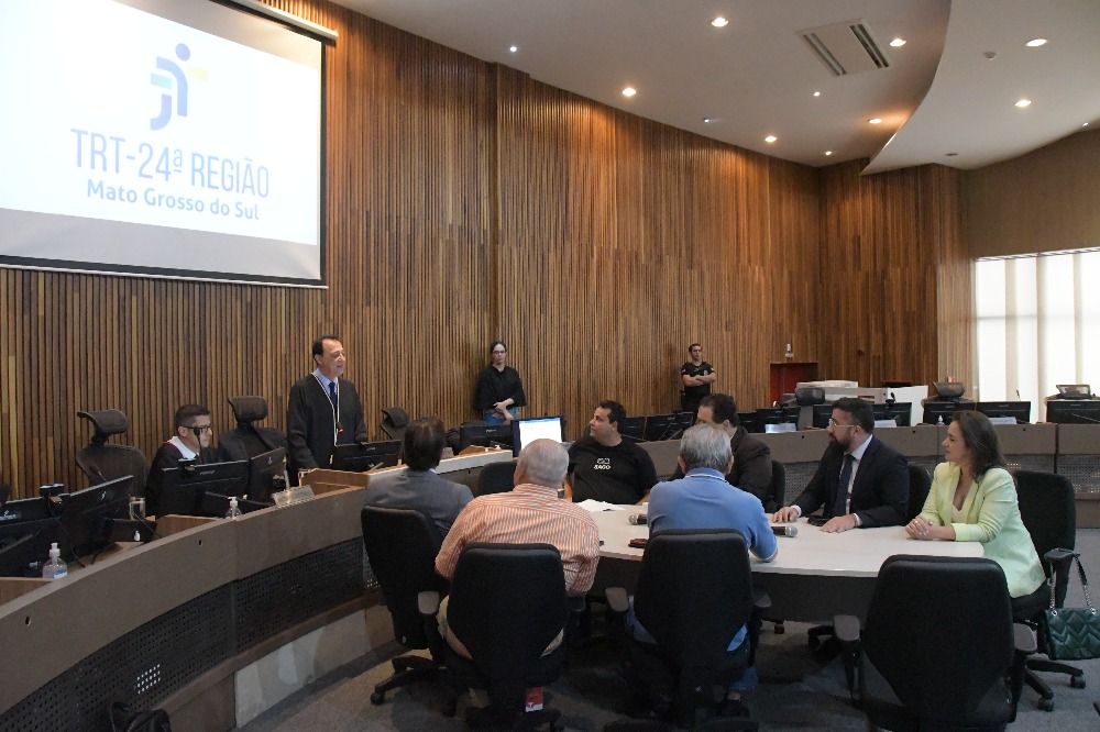 Foto da audiência de conciliação entre os sindicatos do transporte coletivo, a prefeita Adriane Lopes e o Ministério Público do Trabalho. Se reúnem em volta de uma mesa oval os representantes das partes e o desembargador Tomás Bawden está em pé atrás de sua mesa.