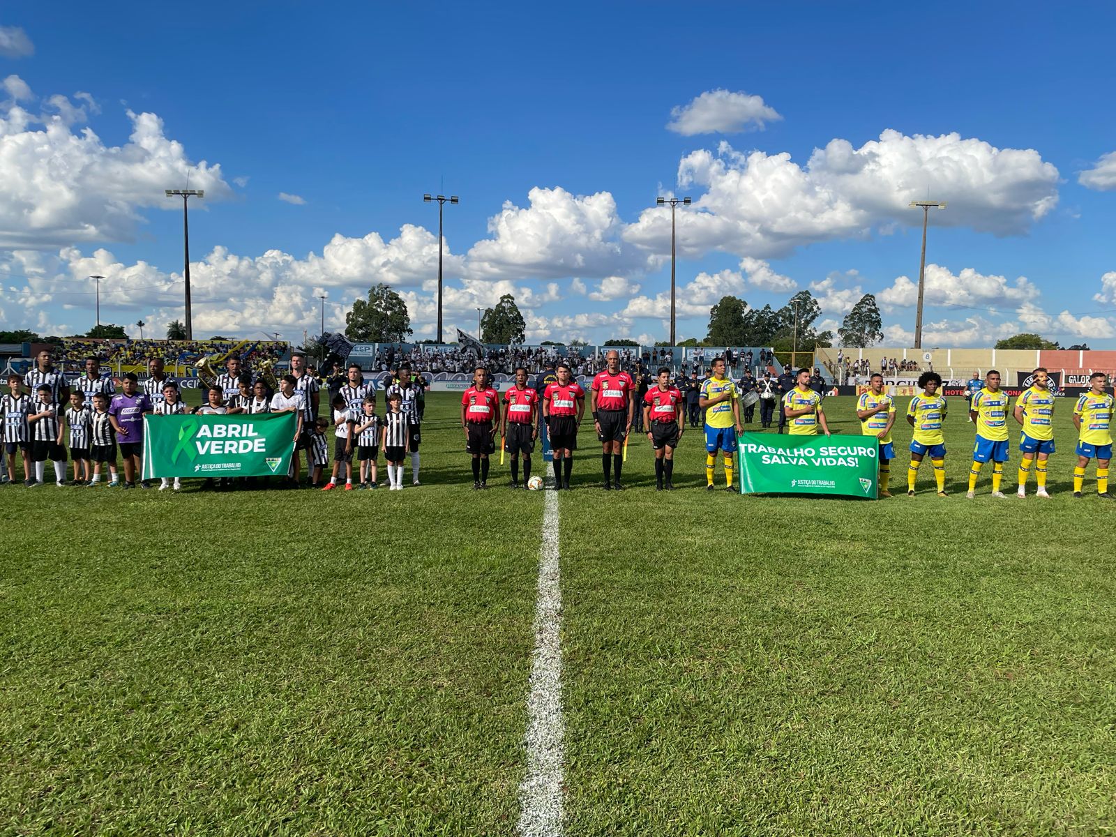 Jogadores de dois times enfileirados no gramado segurando faixas verdes com texto em branco: Abril Verde. Trabalho seguro salva vidas.