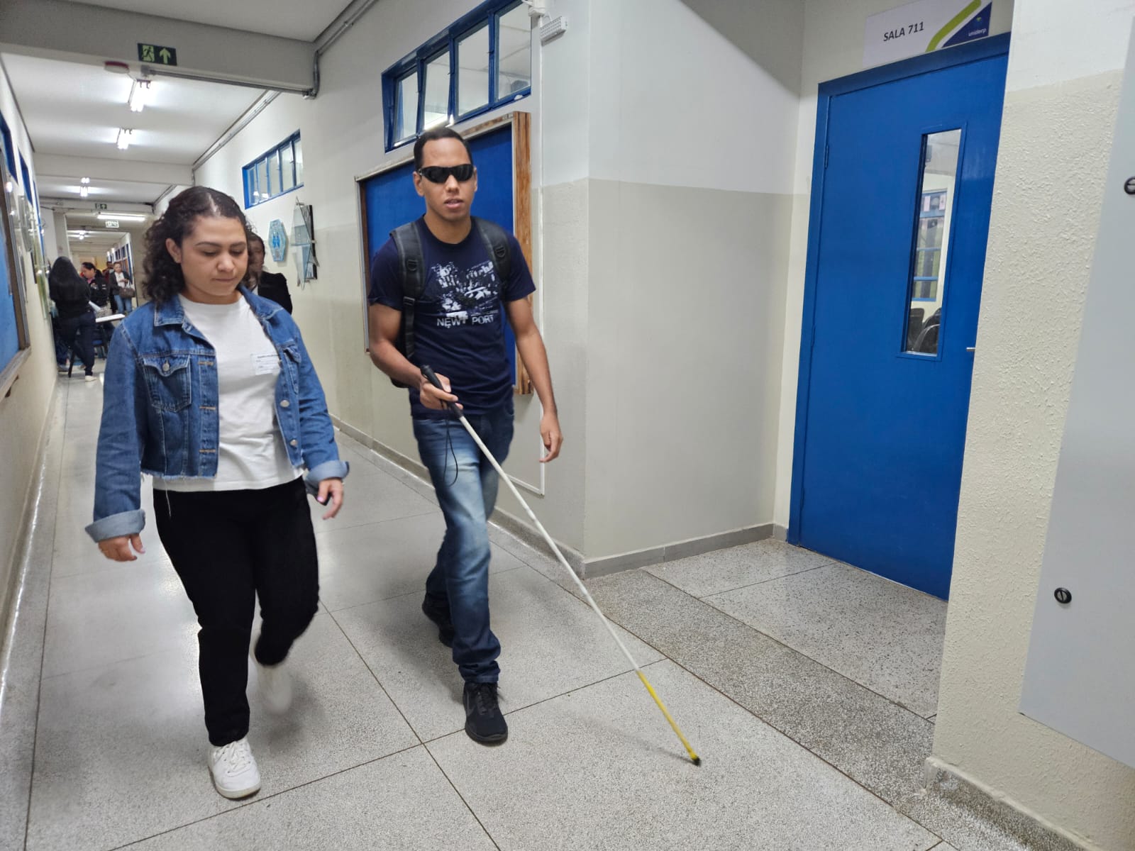 Jovem caminhando com bengala e óculos escuro, acompanhado por mulher
