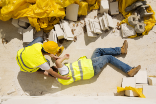 Foto de homem caído no chão com escombros. Ao lado outro homem usando capacete segura as costas do colega.